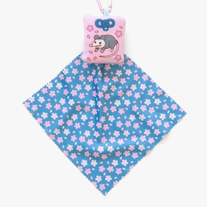 May include: A pink charm with a gray opossum illustration and cherry blossom accents, attached to a blue floral patterned cloth. The cloth has a square shape and is decorated with pink cherry blossoms.