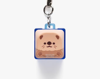 May include: A square-shaped keyring featuring a light brown cartoon otter character with dark brown ears and paws. The otter is set against a blue frame with a light blue handle at the top. The text Geoffrey Siu and itsgeoffrey is printed.