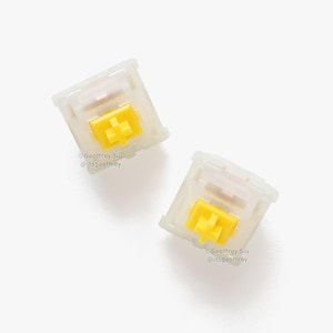 以下が含まれることがあります： 黄色の軸を持つ白い機械式キーボードのキーキャップ2つ。キーキャップは半透明で、わずかに曇りガラスのような仕上げになっています。