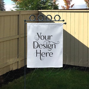 May include: A white garden flag with the text "Your Design Here" in black script is displayed on a black metal stand. The flag is rectangular and hangs in front of a wooden fence and green grass.