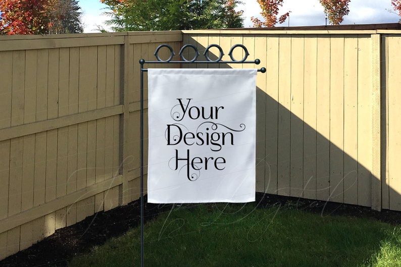 May include: A white garden flag with the text "Your Design Here" in black script is displayed on a black metal stand. The flag is rectangular and hangs in front of a wooden fence and green grass.