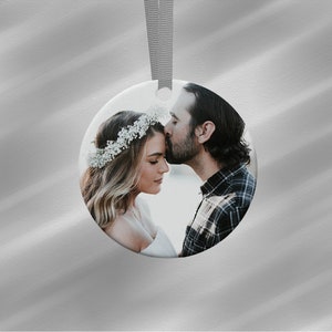 Puede incluir: Un adorno redondo de cer&aacute;mica blanca con una fotograf&iacute;a de una pareja. La mujer lleva una corona de flores y el hombre la besa en la frente. Una cinta gris est&aacute; unida para colgarlo.