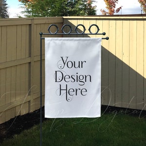 May include: A white garden flag with the text "Your Design Here" in black script hangs from a black metal stand. The flag is displayed outdoors in front of a wooden fence and green grass.
