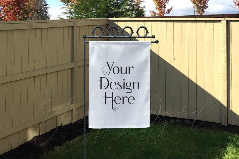 May include: A white garden flag with the text "Your Design Here" in black script hangs from a black metal stand. The flag is displayed outdoors in front of a wooden fence and green grass.