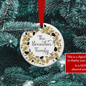K&ouml;nnte beinhalten: Ein wei&szlig;er Keramik-Weihnachtsschmuck mit rotem Band, mit dem Text "The Brandon Family", umgeben von einem goldenen und schwarzen Stechpalmenkranz. Der Schmuck wird an einem Weihnachtsbaum pr&auml;sentiert.