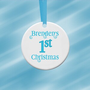 Puede incluir: Adorno navide&ntilde;o de cer&aacute;mica blanca con el texto "Brenden's 1st Christmas" en turquesa. El adorno redondo tiene una cinta turquesa para colgar. El fondo es azul claro.