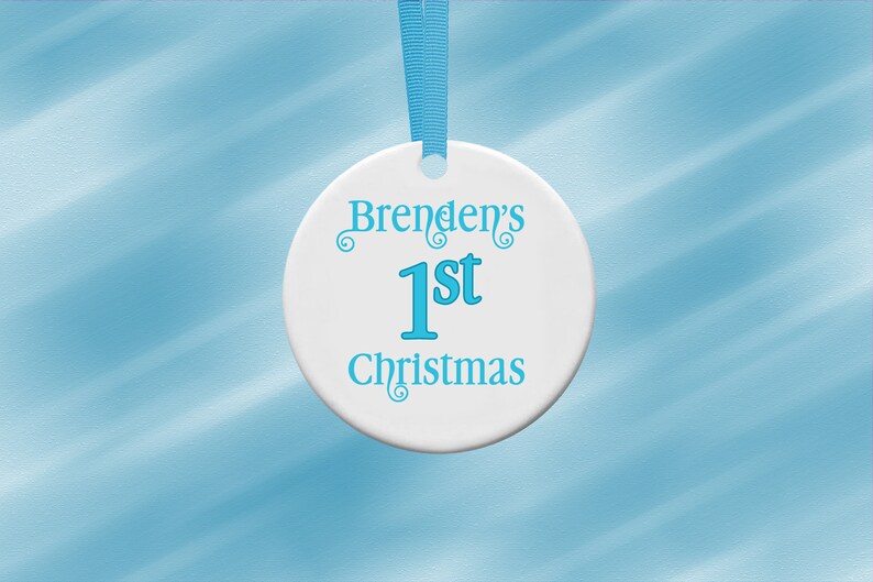 Puede incluir: Adorno navide&ntilde;o de cer&aacute;mica blanca con el texto "Brenden's 1st Christmas" en turquesa. El adorno redondo tiene una cinta turquesa para colgar. El fondo es azul claro.