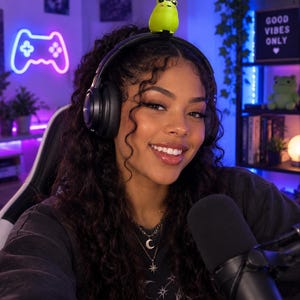 May include: A pair of black over-ear headphones featuring a small, bright green frog figurine attached to the top of the headband. The headphones have a sleek, solid black finish with circular ear cups and a secure, adjustable band design.