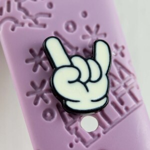 Puede incluir: Una pulsera de reloj de silicona morada con un dije de mano de Mickey Mouse blanco y negro. El dije está en un gesto de mano de rock and roll.