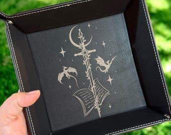 May include: Black square tray with white stitching and a fantasy-themed design. The tray features a central illustration of a sword, crescent moon, open book, stars, and dragons. The tray is held by a hand against a green background.