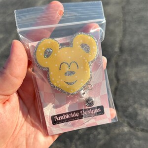 Op de afbeelding: Een gele en zilveren glitter muisvormige intrekbare badgehouder met witte stippen. De badgehouder heeft een zwarte neus en ogen. De tekst "Ambicide Designs" is gedrukt op een roze en witte achtergrond.