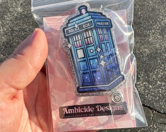 May include: A blue and silver glitter TARDIS charm with the text "POLICE BOX" on the side. The charm is packaged in a clear plastic bag with a pink and white checkered background. The bag also has a sticker that says "Ambicide Designs" and "Locally Crafted for Culture & Style".