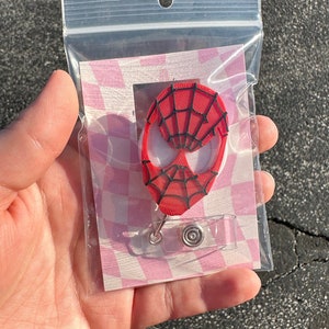 Puede incluir: Un portagafete con un personaje de Spider-Man rojo y negro y un clip de plástico transparente. El portagafete está empaquetado en una bolsa de plástico transparente con un fondo a cuadros rosa y blanco.