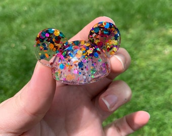 May include: A clear resin ring with a colorful confetti design. The ring is shaped like Mickey Mouse ears.
