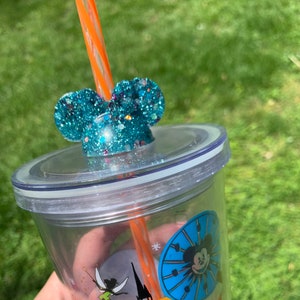 May include: A clear plastic tumbler with a blue and white Mickey Mouse design. The tumbler has a lid with a turquoise glitter Mickey Mouse ear topper and an orange straw.