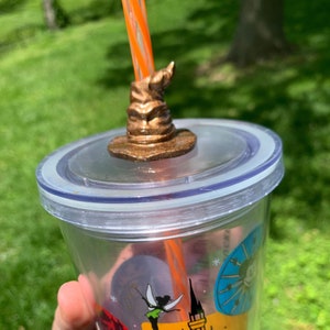 May include: A clear plastic tumbler with a colorful design featuring a castle and a fairy. The tumbler has a lid with a brown Sorting Hat straw topper.