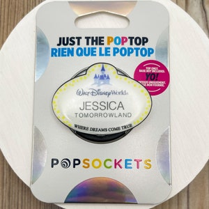 Pode incluir: Um PopSocket branco e amarelo com uma etiqueta de nome do Disney World com o texto "WALT Disney World", "JESSICA", "TOMORROWLAND" e "WHERE DREAMS COME TRUE". O PopSocket é embalado em uma caixa branca e prateada com o texto "JUST THE POPTOP", "RIEN QUE LE POPTOP" e "POPSOCKETS".