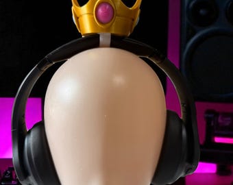 May include: A mannequin head wearing black headphones and a gold crown with a pink gem. The background includes a computer monitor and speakers, suggesting a gaming or audio setup.