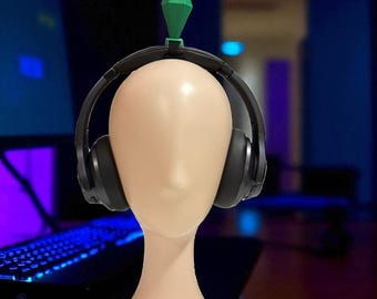 May include: A mannequin head with black headphones and a green diamond-shaped object on top. The mannequin is on a wooden surface, with a computer and keyboard in the background. The scene is lit with blue and purple hues.
