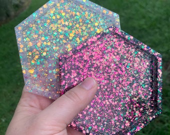 May include: Two hexagonal resin coasters. One is clear with iridescent glitter, the other is black with pink and iridescent glitter. The coasters are held up against a green background.