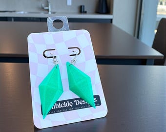 May include: A pair of bright green, geometric earrings with a diamond shape, hanging from silver hooks. The earrings are displayed on a pink and white checkered card with the text "Ambicide Designs" and "ALLY CRAFTED POP-CULTURE TREASURES".