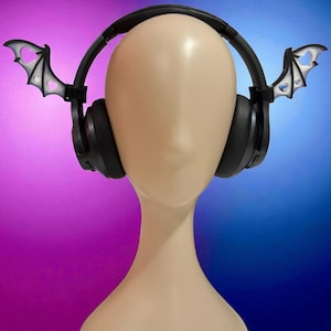 May include: Black headphones with bat wing attachments. The bat wings are white with black outlines and a heart-shaped cutout. The headphones are on a mannequin head against a gradient purple and blue background.