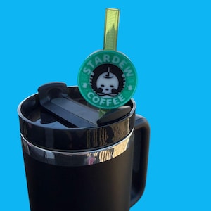 Könnte beinhalten: Schwarzer Reisebecher mit silbernem Rand und einem grün-weißen "Stardew Coffee" Strohhalm-Aufsatz. Der Aufsatz zeigt ein Totenkopf-Design. Der Becher befindet sich auf einem hellblauen Hintergrund.