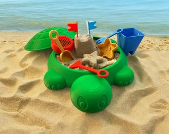 Puede incluir: Una caja de arena con forma de tortuga verde con tapa. La caja de arena está llena de arena e incluye una pala roja, un rastrillo rojo, un cubo azul, un molde en forma de estrella amarilla y dos banderas.