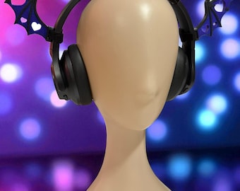 May include: Black headphones with bat wing accents, displayed on a mannequin head. The bat wings are dark blue with heart-shaped cutouts. The background is a blurred mix of purple and blue lights.