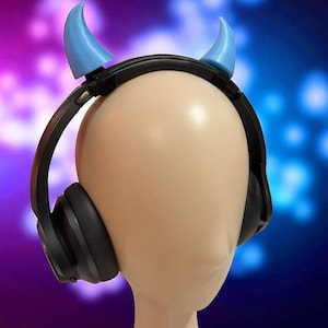 May include: Black over-ear headphones with light blue devil horns attached to the headband. The headphones are on a mannequin head with a neutral skin tone. The background is a blurred mix of purple and blue.