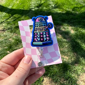 Könnte beinhalten: Ein blauer und lilafarbener Cartoon-Dalek mit glitzerndem Design, befestigt an einem durchsichtigen, einziehbaren Ausweishalter mit einem silbernen Clip.