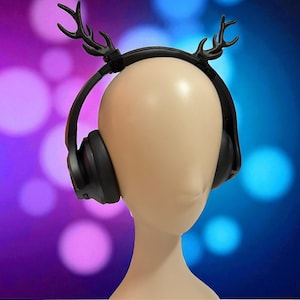 Può includere: Cuffie nere over-ear con corna decorative nere sono esposte su una testa di manichino. Le cuffie sono posizionate sulla testa, con le corna che si estendono verso l'alto. Lo sfondo presenta una sfocatura di tonalità viola e blu.