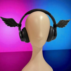 May include: Black over-ear headphones with decorative black wings attached. The headphones are displayed on a mannequin head against a pink and blue gradient background.