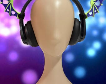 May include: Black over-ear headphones with bat wing accents. The bat wings are lime green with purple and white heart details. The headphones are displayed on a mannequin head against a blurred purple and blue background.