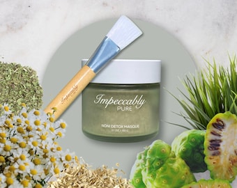 Könnte beinhalten: Ein grünes Glas Noni Detox Masque von Impeccably Pure, mit einem Holz- und weißen Pinsel, umgeben von grünen Blättern, weißen Blüten und getrockneten Kräutern.