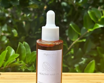 Könnte beinhalten: Eine braune Glasflasche mit einer weißen Tropfspitze und einem weißen Etikett. Das Etikett lautet "Impeccably Pure" und "Healthy Hair Oil". Das Etikett lautet auch "Strengthen | Lengthen | Repair" und "Organic Ingredients".