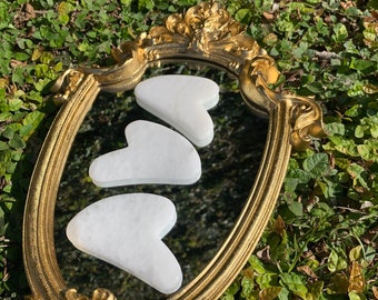 Könnte beinhalten: Drei weiße herzförmige Gua Sha-Werkzeuge werden in einem goldenen ovalen Spiegelrahmen präsentiert. Die Werkzeuge sind aus glattem, poliertem Stein gefertigt und werden übereinander gestapelt.