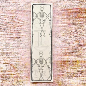 May include: A rectangular bookmark with a black border and a repeating pattern of skeleton illustrations. The skeletons are depicted in a simple, line-art style, with arms crossed. The background is a light beige colour.