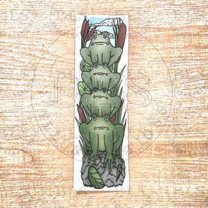 Könnte beinhalten: Ein rechteckiges Lesezeichen mit einem vertikalen Stapel aus vier grünen Fröschen, die auf einem grauen Felsen sitzen. Die Illustration zeigt hohe grüne Schilfhalme, braune Rohrkolben und Seerosen vor einem hellblauen Himmel.