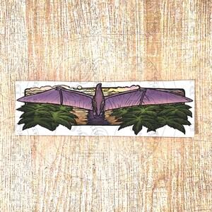 Puede incluir: Una pegatina rectangular que presenta una ilustración detallada de un pterodáctilo morado con las alas extendidas. La criatura está situada sobre un río oscuro rodeado de un denso follaje verde bajo un cielo de color amarillo pálido y naranja.