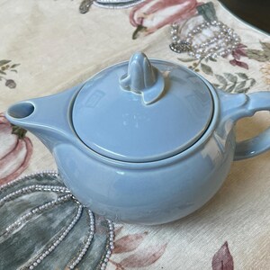 May include: A light blue ceramic teapot with a matching lid. The teapot has a curved handle and a spout. The lid features a decorative finial. The teapot is sitting on a floral patterned tablecloth.