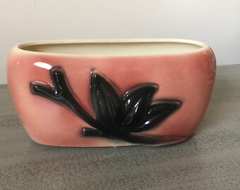 May include: A pink ceramic planter with a black floral design. The planter is oval shaped and has a glossy finish.