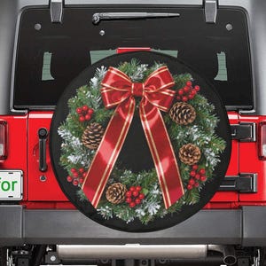 May include: A black spare tire cover featuring a Christmas wreath design with a red bow, pine cones, and berries. The cover is mounted on the back of a red Jeep. The license plate reads "Amfor".
