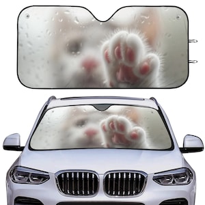 Op de afbeelding: Een witte voorruitzonnescherm met een witte kattenpoot die door een wazig raam reikt. De zonnekap is ontworpen om te voorkomen dat de zon en de warmte de auto in komen.