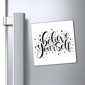 May include: White square refrigerator magnet with black lettering that reads "Believe in Yourself" with small black stars scattered around the text.