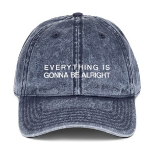 May include: A blue denim baseball cap with the text "EVERYTHING IS GONNA BE ALRIGHT" embroidered in white on the front.