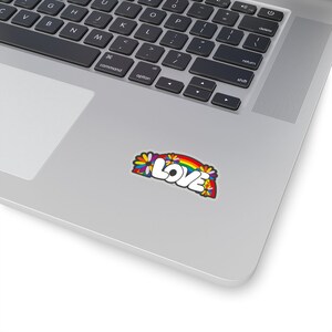 May include: A sticker with a rainbow arch and the word "LOVE" in white letters with a black outline. The rainbow is made up of the colors red, orange, yellow, green, blue, indigo, and violet. There are flowers on either side of the rainbow.