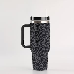 Könnte beinhalten: Ein hoher, isolierter Reisebecher mit schwarzem Griff und Deckel. Der Becher hat ein graues Leopardenmuster und einen durchsichtigen Strohhalm. Der Edelstahlrand und der Griff bilden einen eleganten Kontrast zum Animal-Print-Design.
