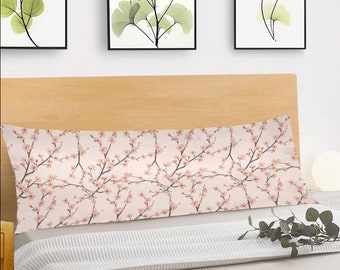 Puede incluir: Una almohada larga y rectangular con un estampado floral rosa y blanco con delicadas flores de cerezo. La almohada está sobre una cama con una manta a rayas blancas y grises.