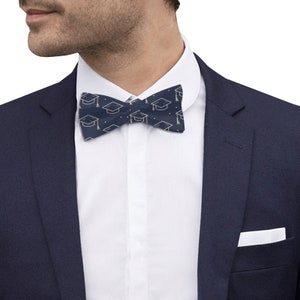 May include: A navy blue bow tie with a gold graduation cap pattern. The bow tie is tied around a white collared shirt.